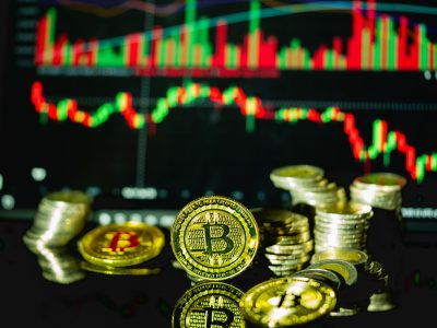 Bitcoin ve metal sikke yığınları, arkada kırmızı-yeşil mum grafiği