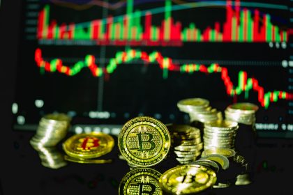 Bitcoin ve metal sikke yığınları, arkada kırmızı-yeşil mum grafiği
